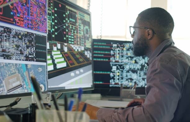 Engineer using computer-aided engineering tools to analyze circuit board layouts and electronic system simulations across multiple screens.