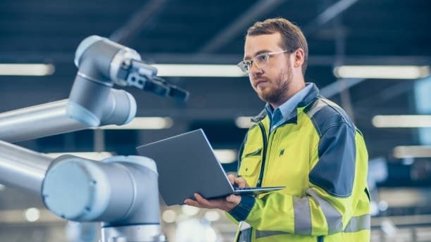 Engineer programming a robotic arm with a laptop in an automated CNC robotics environment