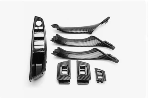 A set of black injection-molded plastic components featuring a carbon-fiber texture, including trim panels and switch bezels